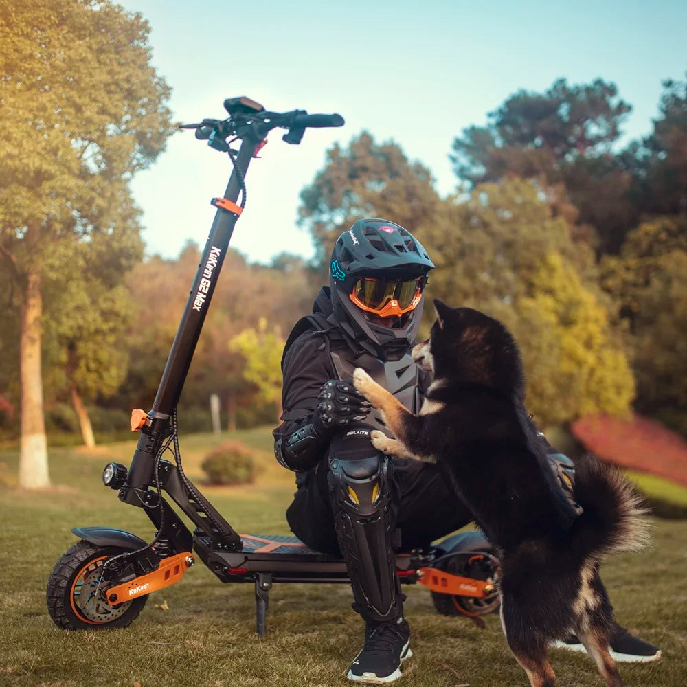 A rider with protective gear interacts with his dog while seated on the black and orange KuKirin G2 Max off-road electric scooter in a sunny park.​